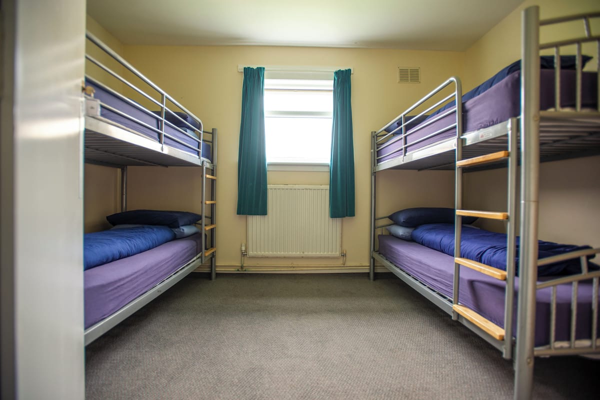 A dormitory room with bunk beds, a window with curtains, and a radiator.