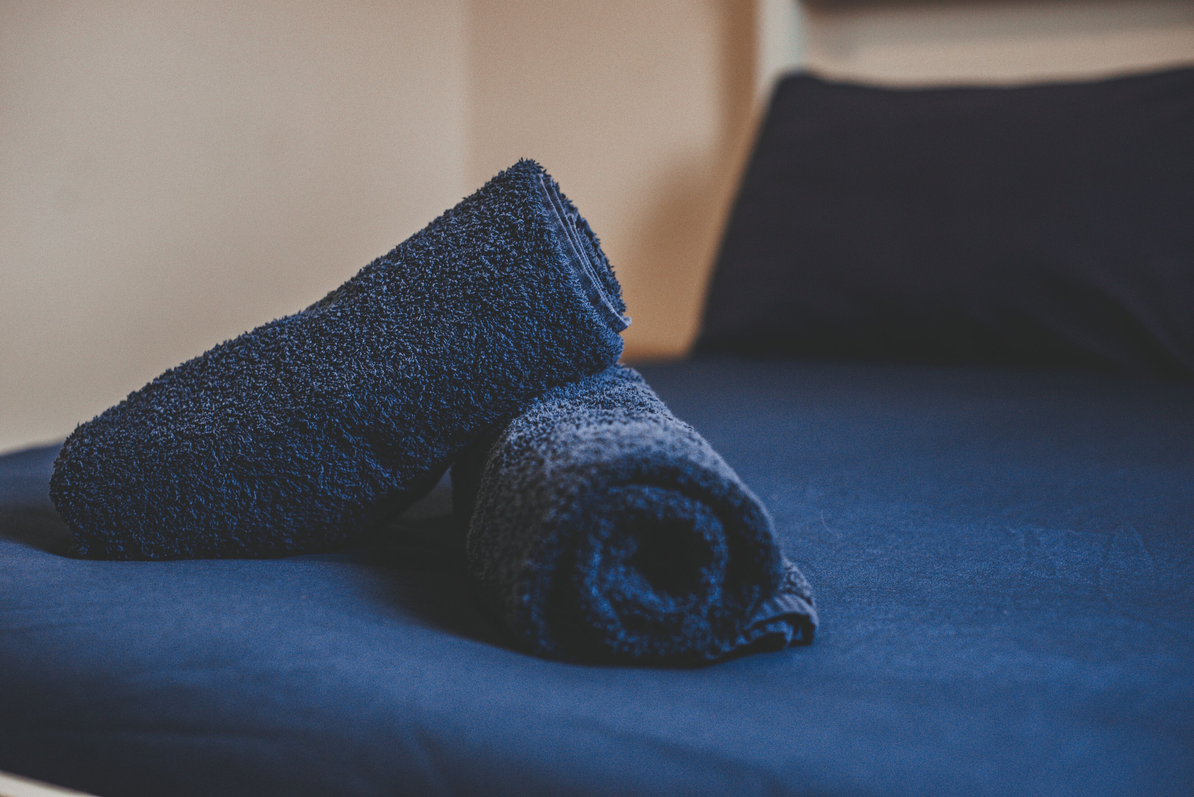 A neatly made bed with a dark blue bedspread and two rolled-up towels.