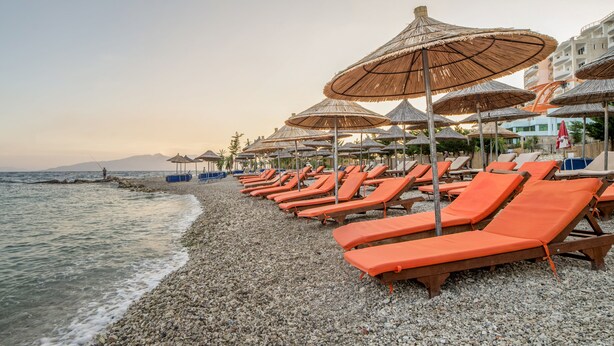 A beach with rows of orange lounge chairs and thatched umbrellas, a pebbled beach, and a view of the sea and distant mountains. - Vila Kalcuni (Sarandë)