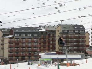 A ski lift with a person in a chairlift. There are multi-story buildings with balconies in the background.