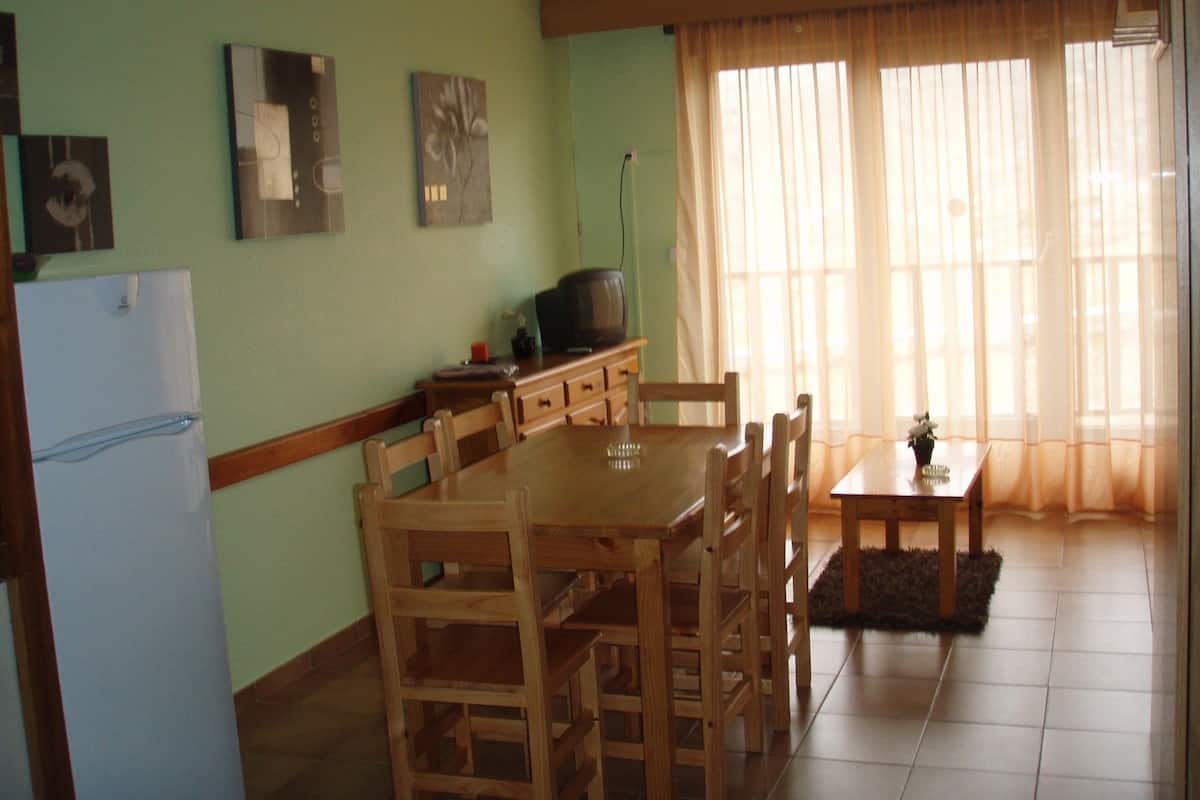 A dining area with a wooden table and chairs, a television on a stand, and a refrigerator.