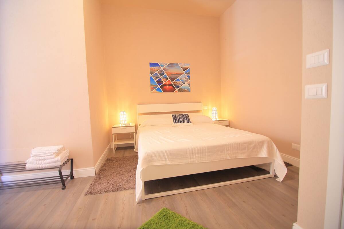 A neatly arranged bedroom with a bed, bedside lamps, a wall-mounted artwork, and a towel rack.
