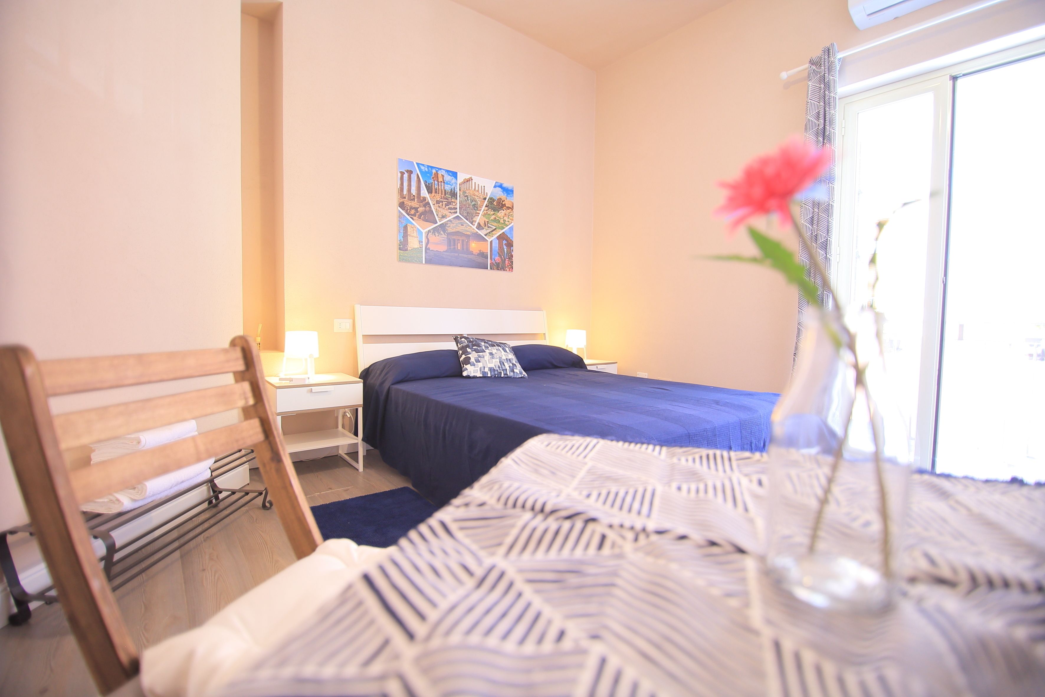 A bedroom with a bed, a nightstand, a lamp, a window with curtains, and a wall with a picture.