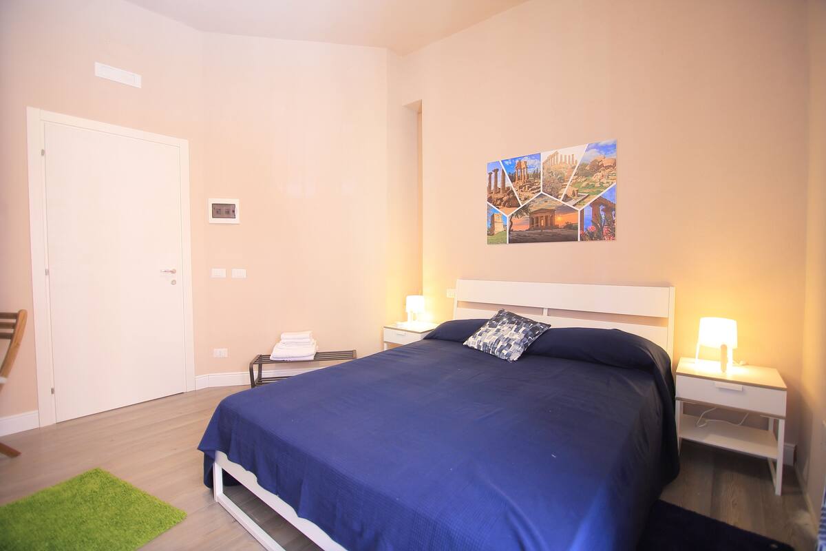 A bedroom with a large bed, two bedside tables with lamps, a green rug, and a wall art piece.