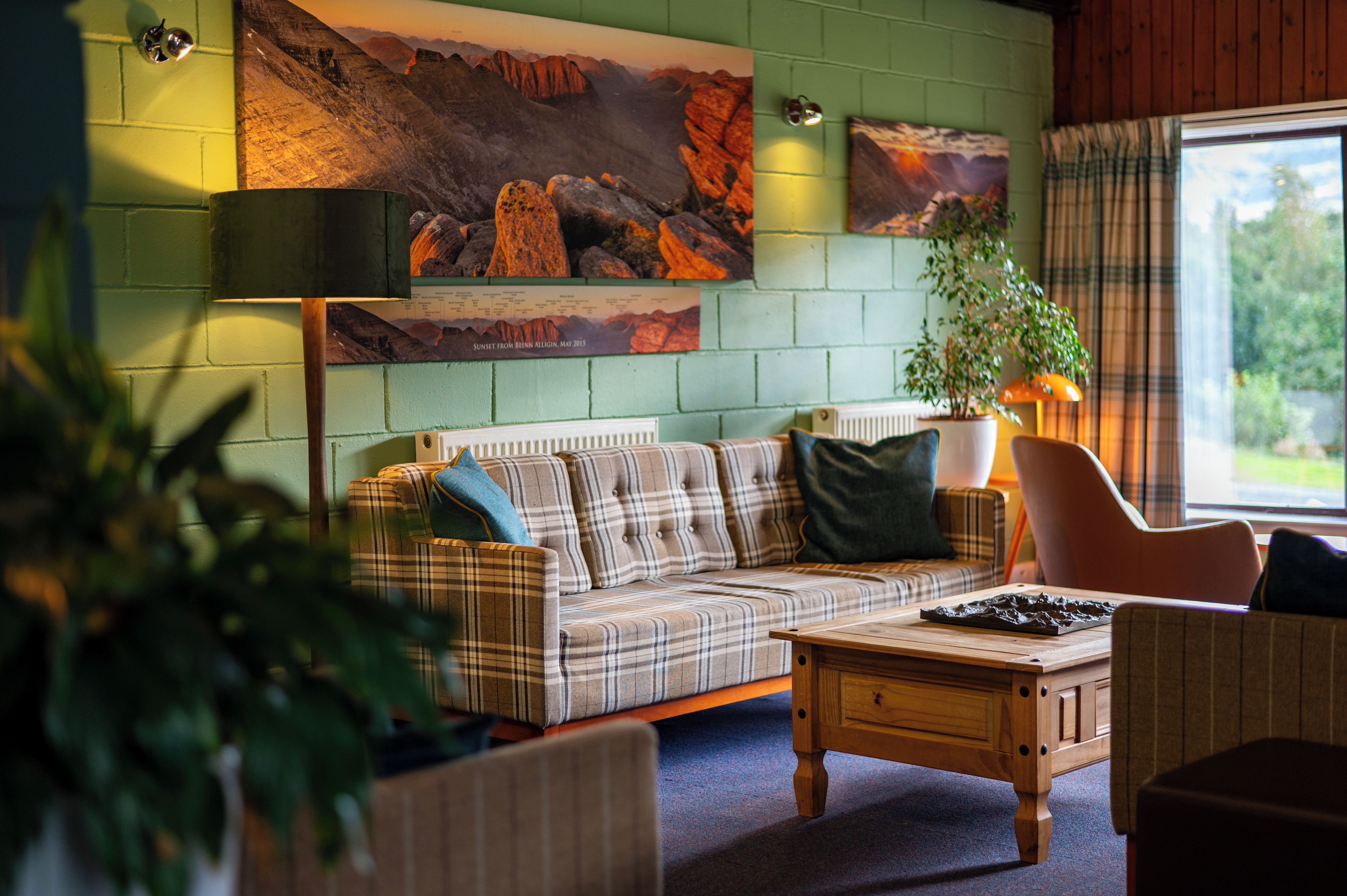 A room with a plaid sofa, a wooden coffee table, and a chair. There are two wall-mounted lamps and a large painting of a mountain landscape.