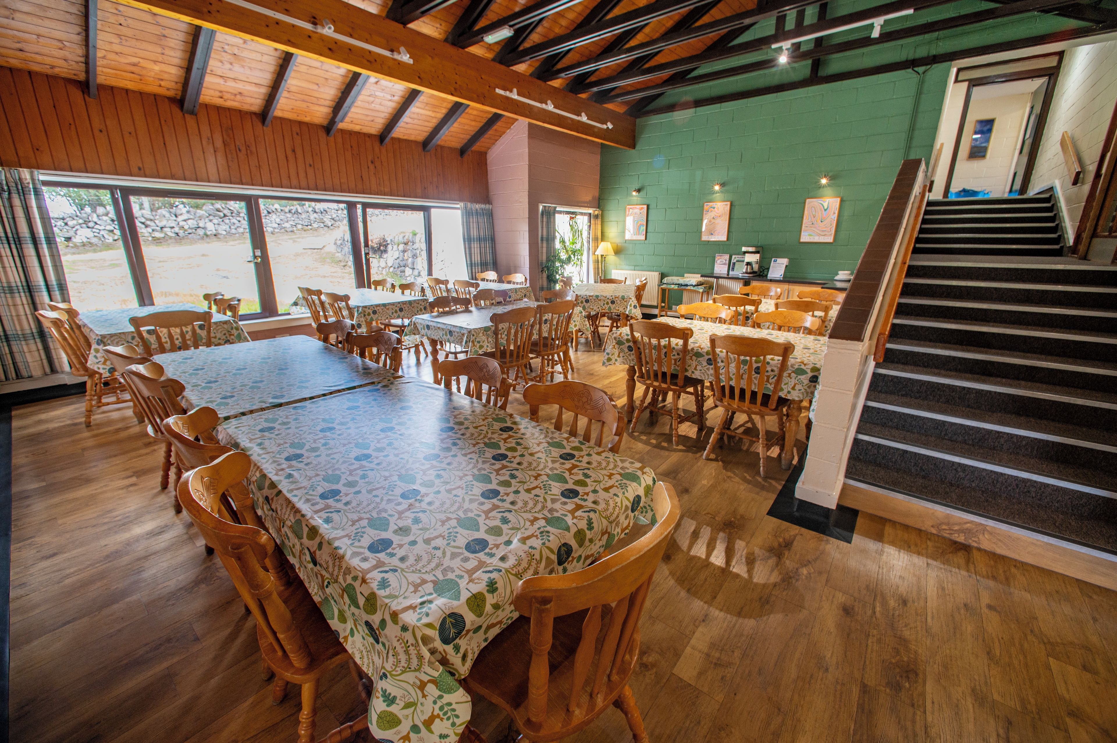 A restaurant with wooden tables and chairs, a green wall, and a staircase leading to an upper level.