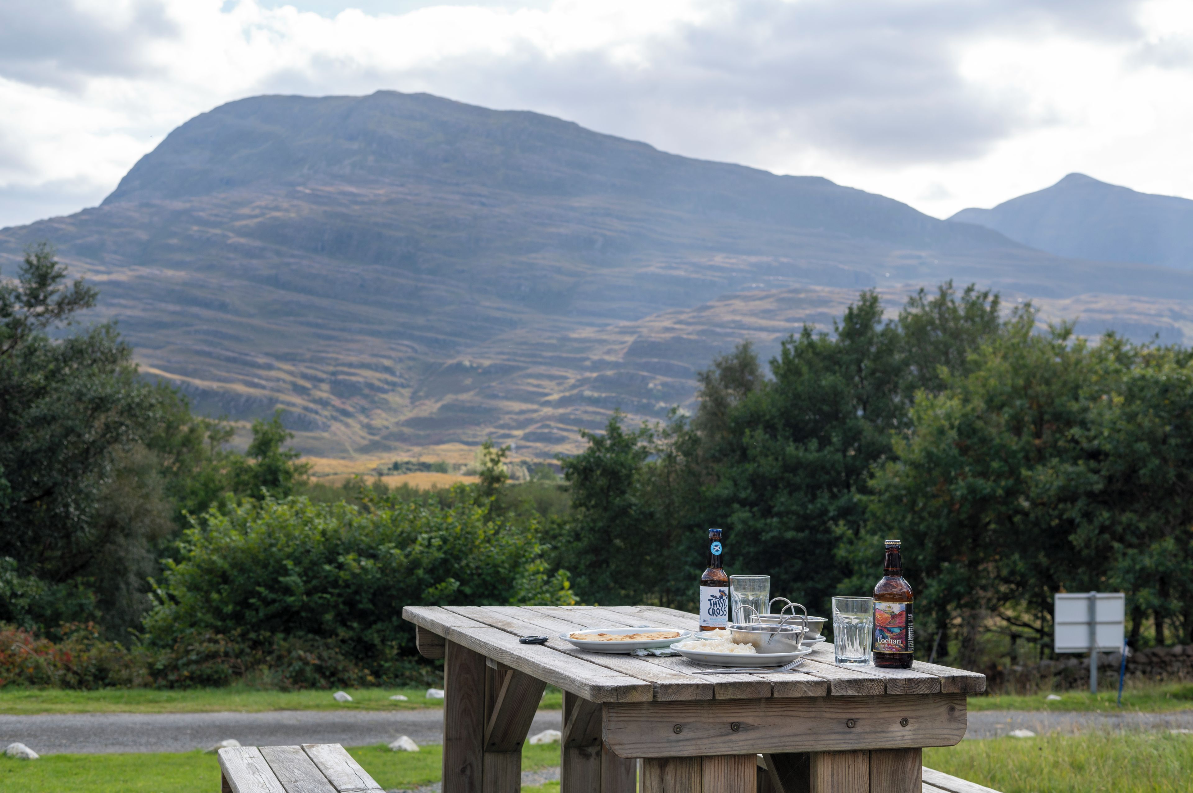 Une table de pique-nique avec des assiettes, des verres et des bouteilles, le tout installé dans un cadre naturel avec des montagnes en arrière-plan.