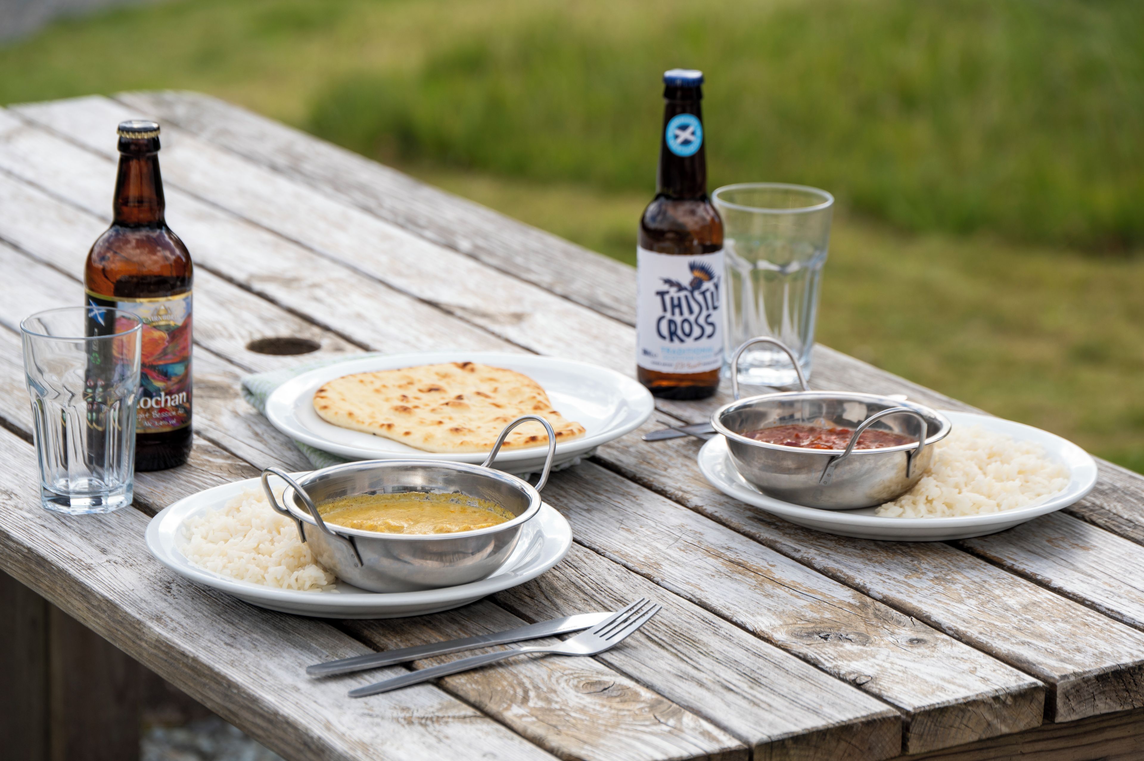 A picnic setup on a wooden table with plates of food, a bottle of beer, and a glass.