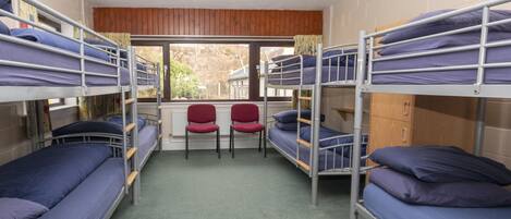 A dormitory room with bunk beds, a radiator, a window, and two red chairs.