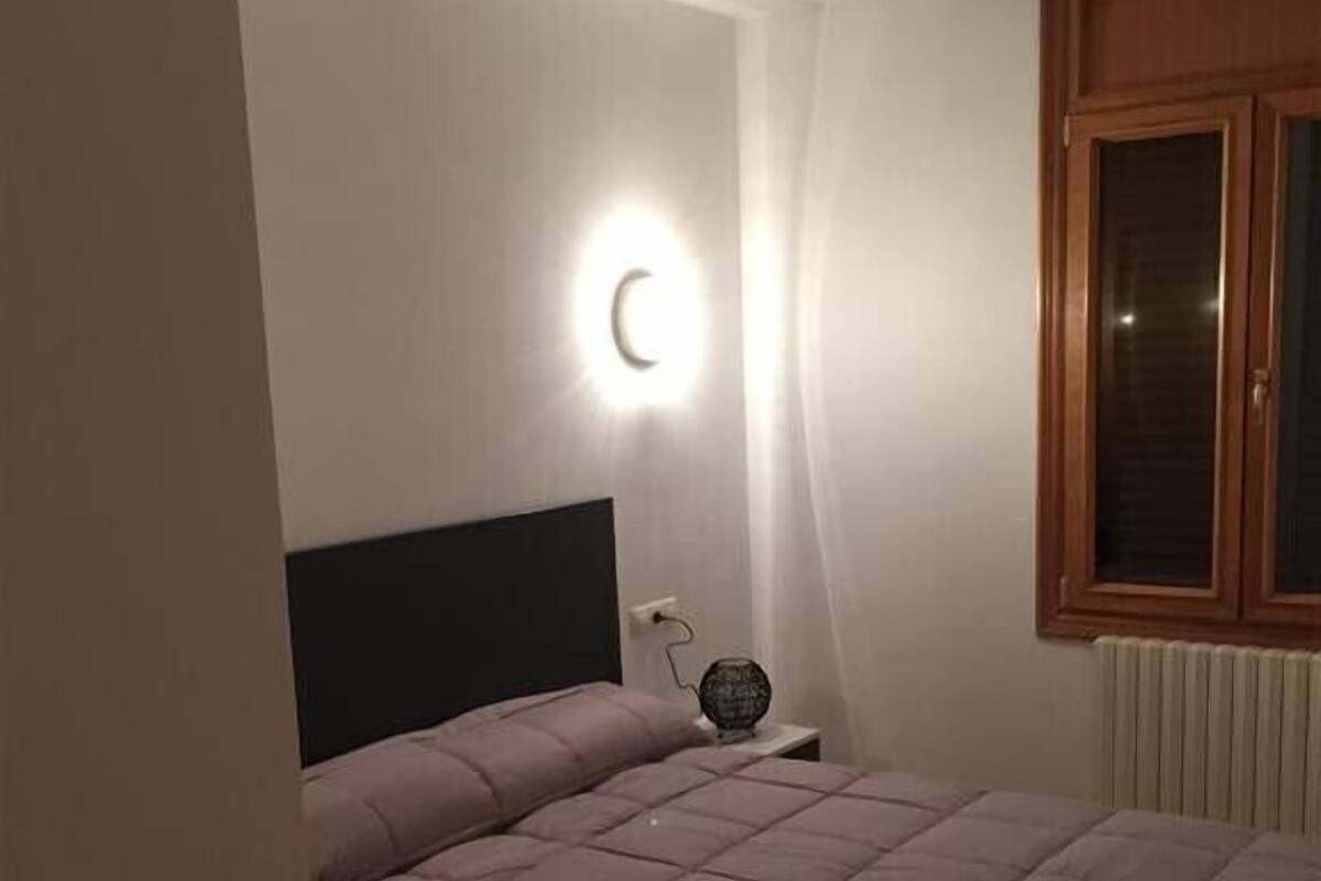 Un dormitorio con cama, mesita de noche, lámpara de pared y ventana con marco de madera.