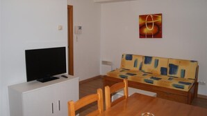 Een kamer met een televisie op een witte kast, een houten tafel met stoelen, en een bank met een kleurrijk abstract schilderij aan de muur.