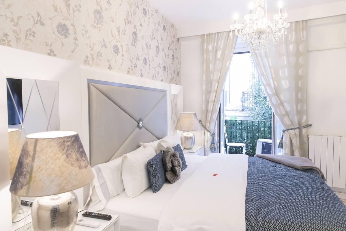 Ein Hotelzimmer mit einem großen Bett, einem Nachttisch mit Lampe, einem Fenster mit Vorhängen und Blick auf Grünflächen.