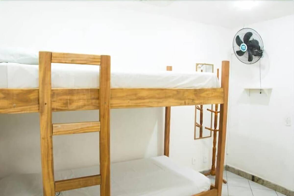 A bunk bed with a wooden frame and white bedding. There is a wall-mounted fan on the right side.