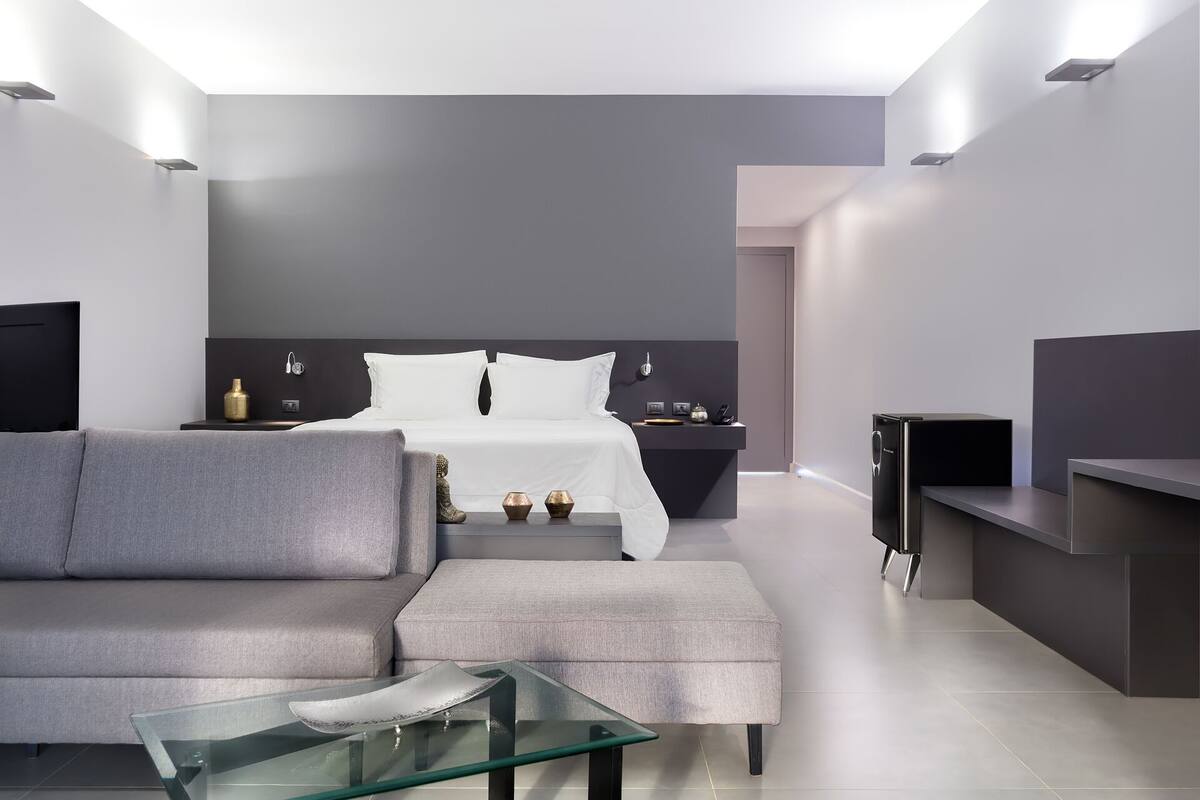 Una habitación de hotel moderna con un sofá gris, una mesa de centro de vidrio, una cama con ropa de cama blanca, una mesita de noche con una lámpara y un televisor sobre un mueble.
