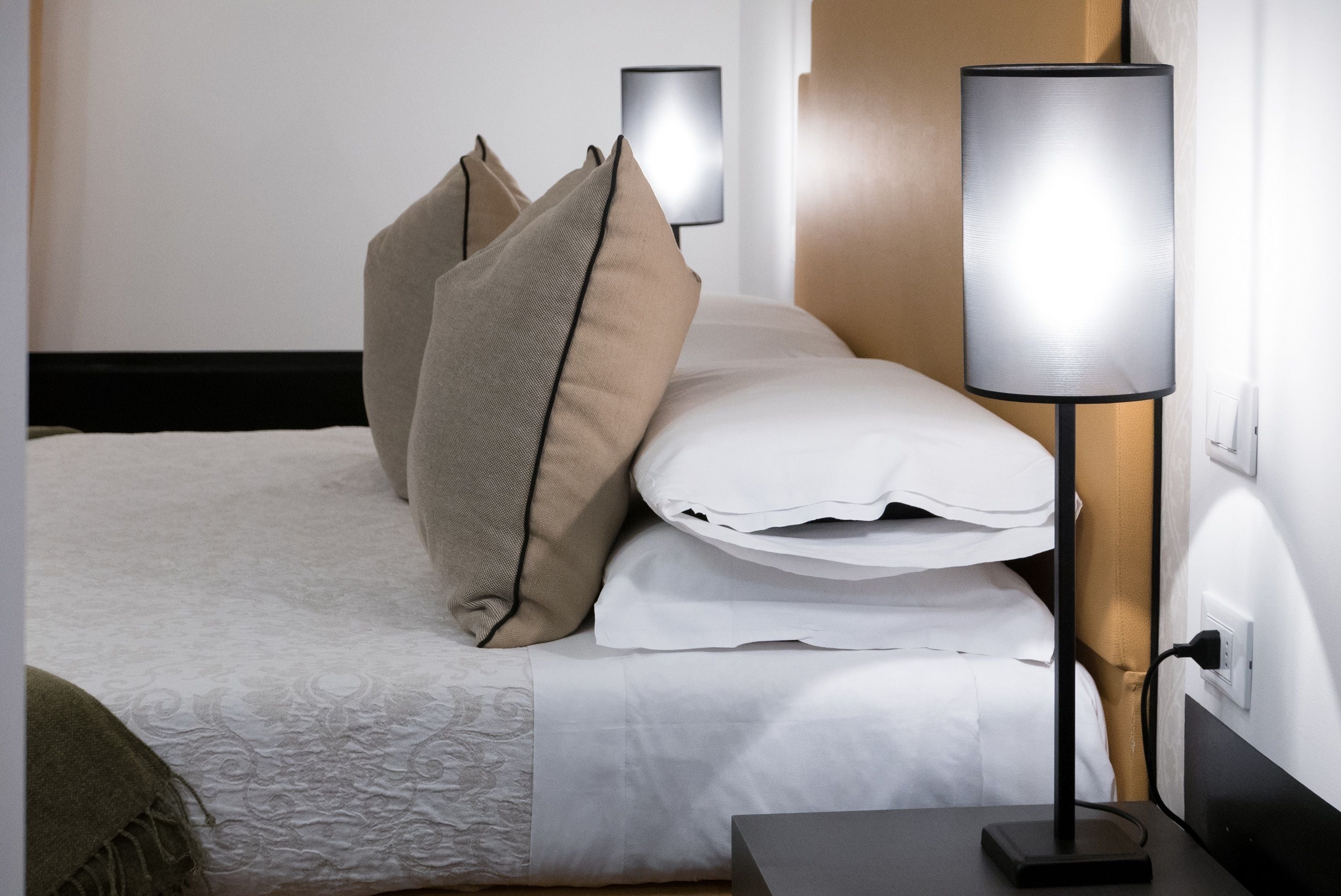 Una cama bien hecha con sábanas blancas y almohadas color beige, una lámpara de noche y un enchufe en la pared.