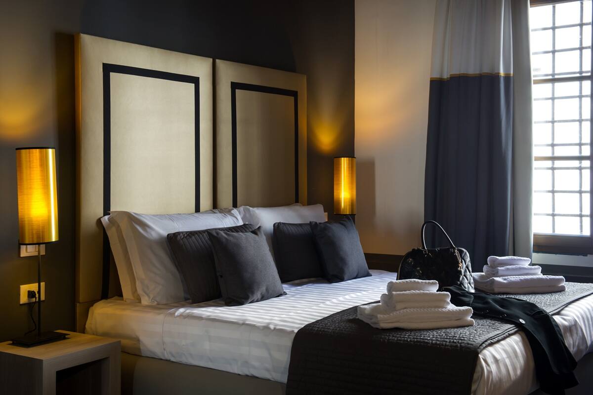 Camera d'albergo con un letto grande, due lampade sui comodini, un comodino, una finestra con tende e un mucchio di asciugamani sul letto.