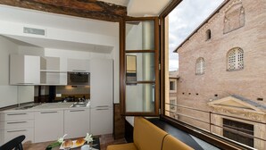 Une cuisine moderne avec des meubles blancs et des appareils en acier inoxydable, un coin repas avec un canapé jaune, et une vue sur un bâtiment historique en briques, visible à travers une grande fenêtre.