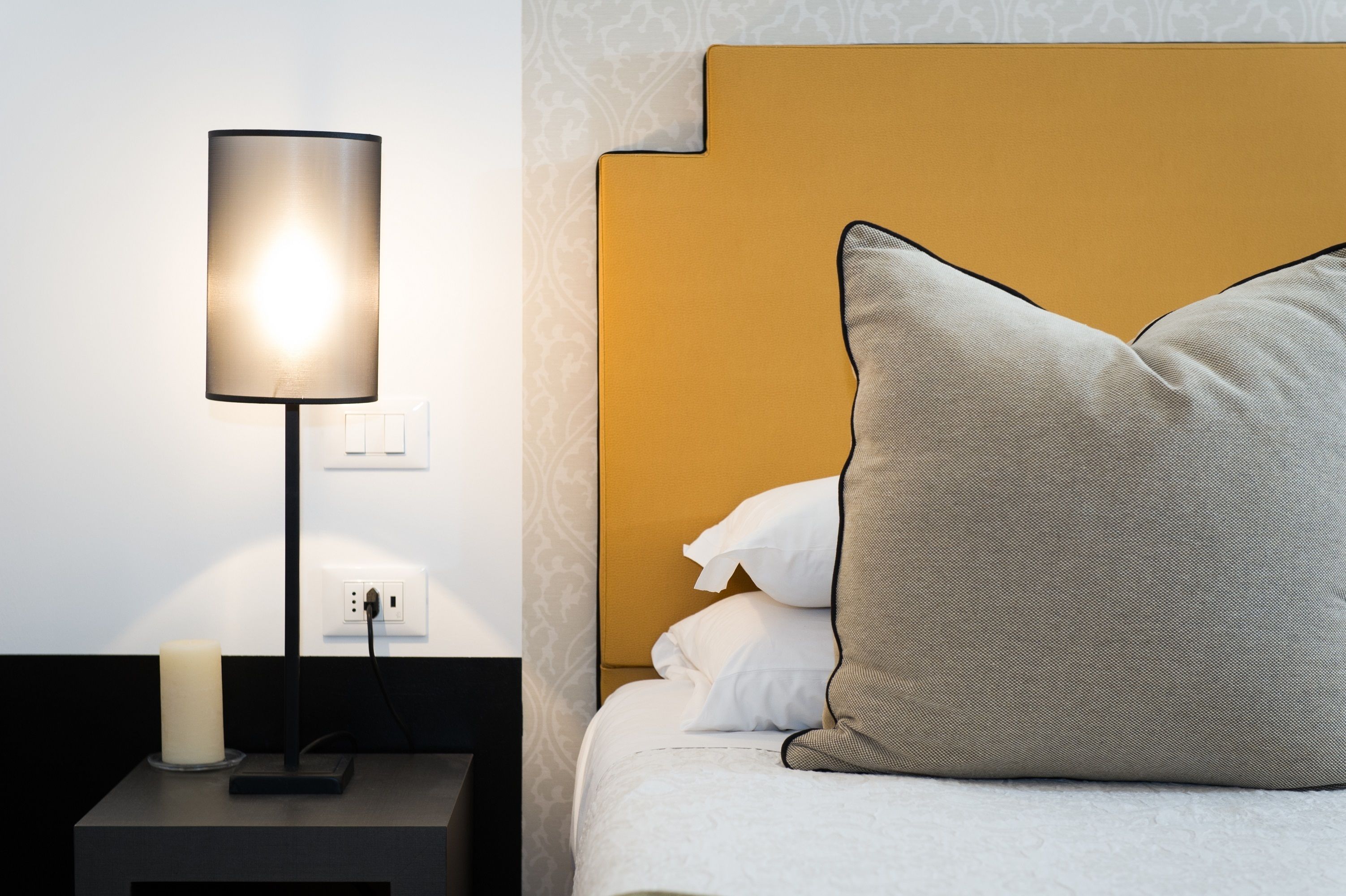 Een netjes ingerichte hotelkamer met een bedlampje, een wit bed met een gestructureerd hoofdeinde en een klein nachtkastje met een kaars.