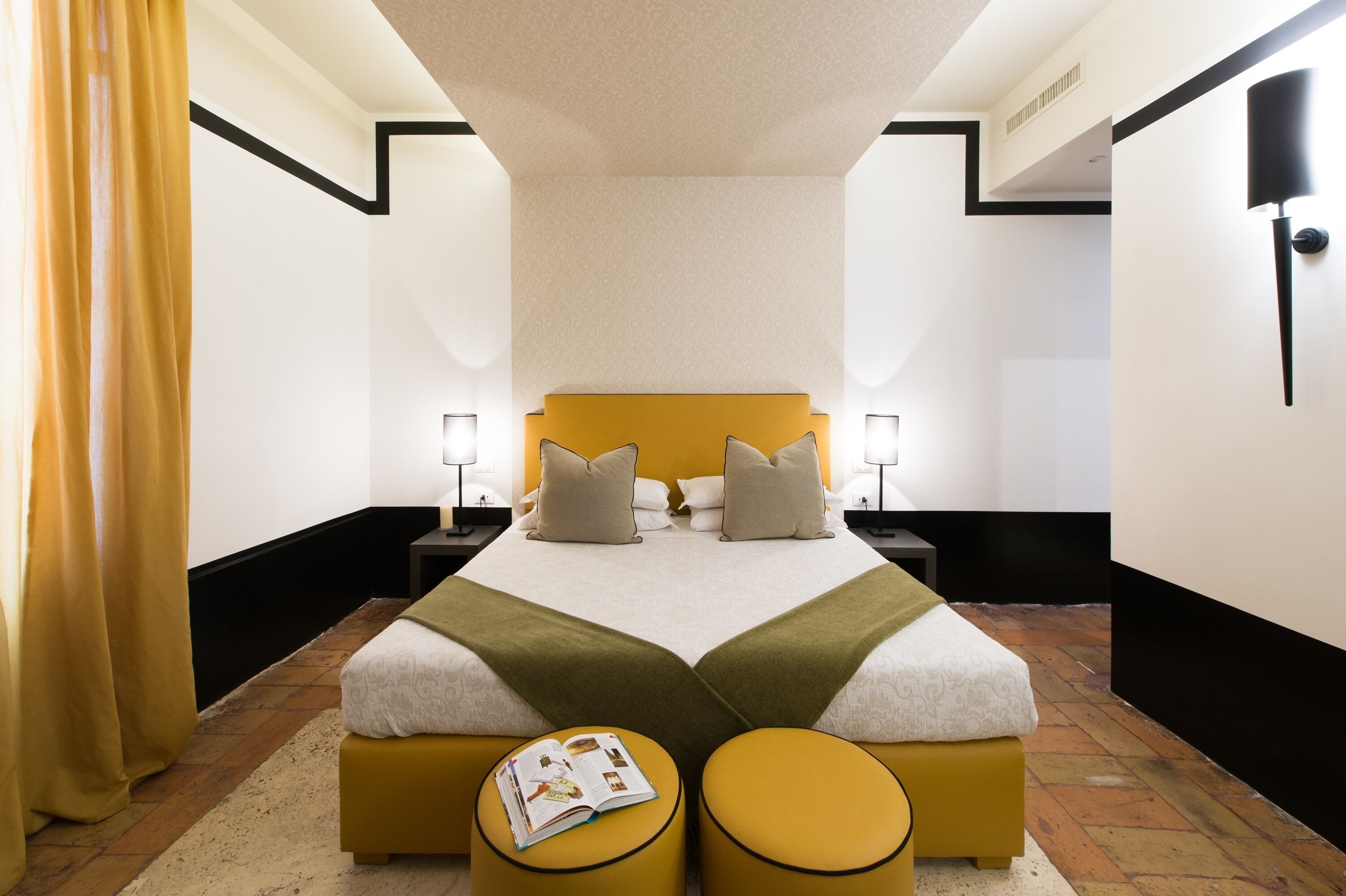 Camera d'hotel con un letto matrimoniale, due comodini con lampade, una testiera gialla e un piccolo tavolino da lettura con una rivista.