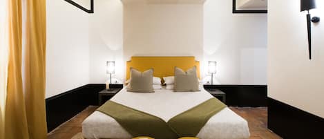 Habitación de hotel con cama doble, dos mesitas de noche con lámparas, cabecera amarilla y una pequeña mesa de lectura con una revista.