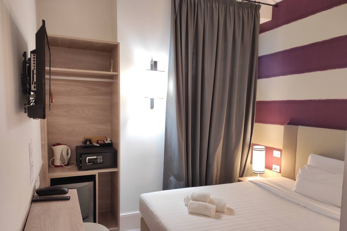 Una camera d'albergo con un letto, una scrivania con una lampada, una TV e un armadio.