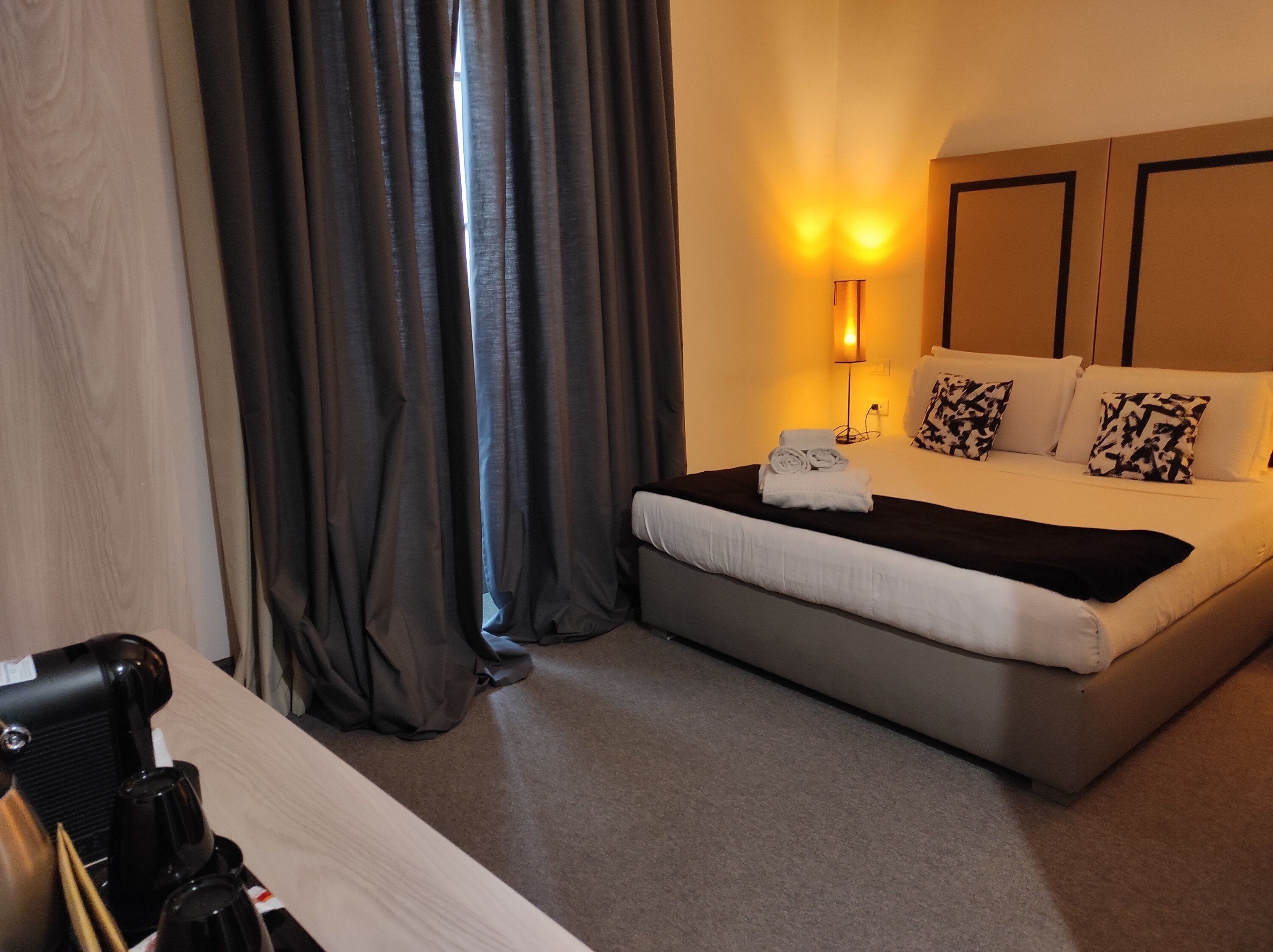 A hotel room with a bed, a bedside table with a coffee maker, a lamp, and a window with curtains.