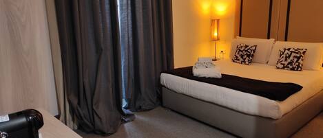 A hotel room with a bed, a bedside table with a coffee maker, a lamp, and a window with curtains.