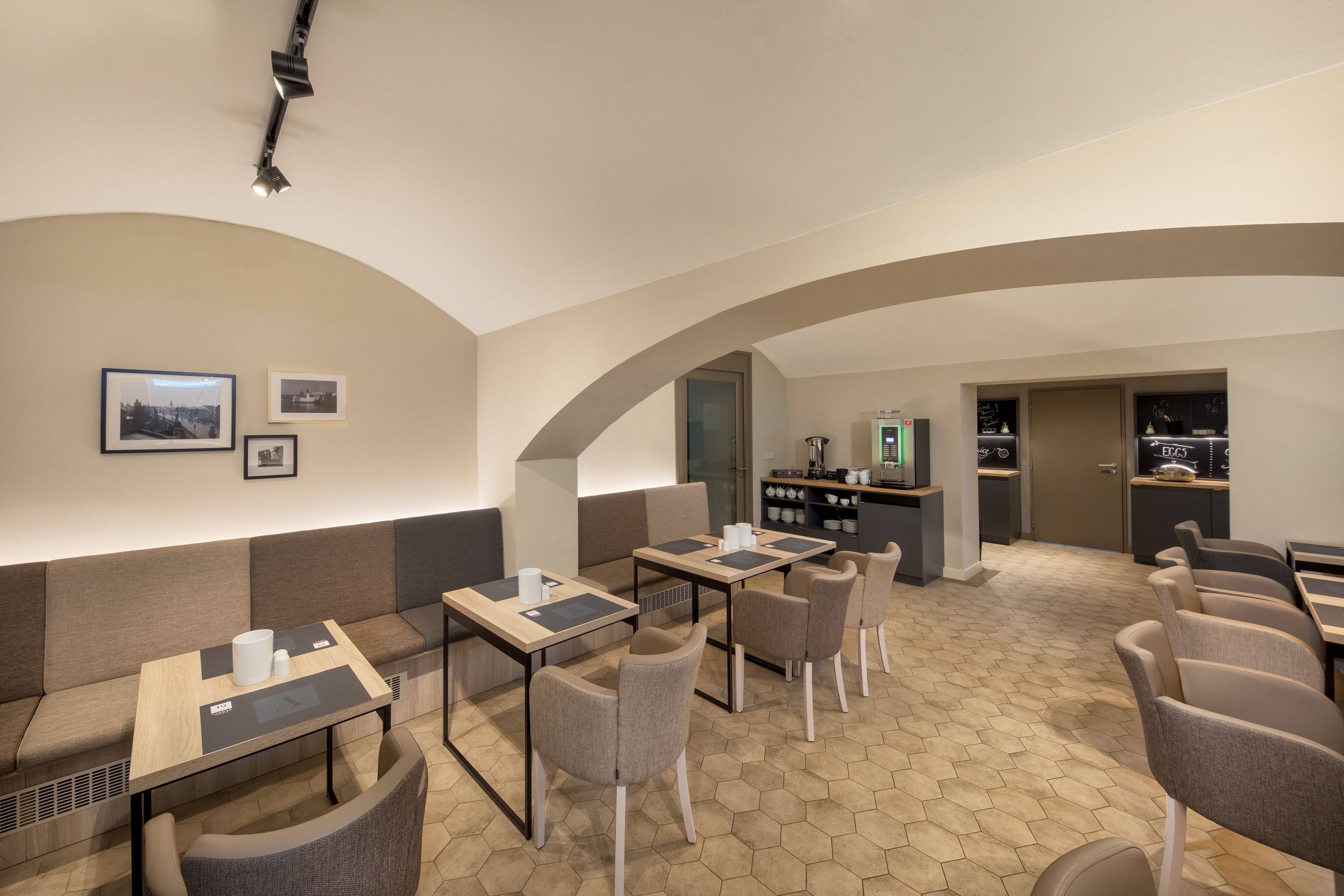 Un café moderno con suelo de patrón hexagonal, mesas de madera con individuales negros y sillas tapizadas en gris.