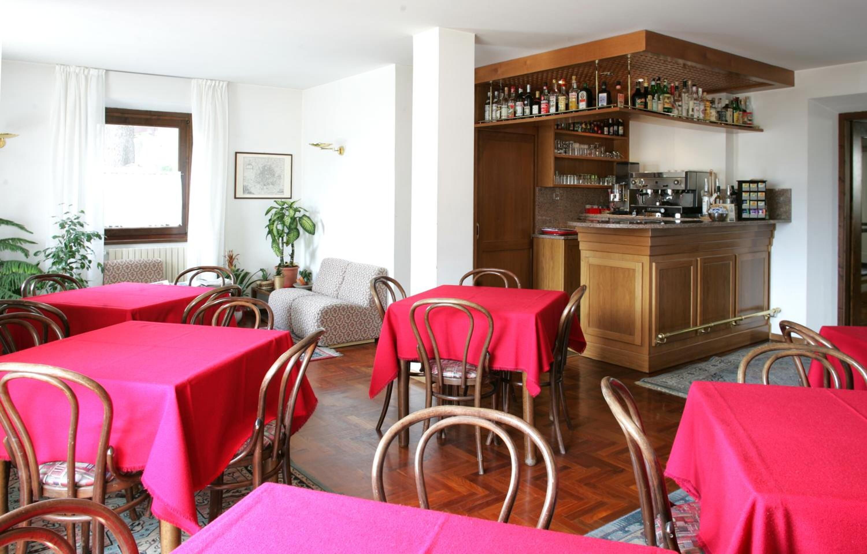 A dining area with red tablecloths and metal chairs, a bar area with shelves of bottles, and a sofa in the background.