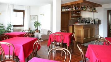A dining area with red tablecloths and metal chairs, a bar area with shelves of bottles, and a sofa in the background.