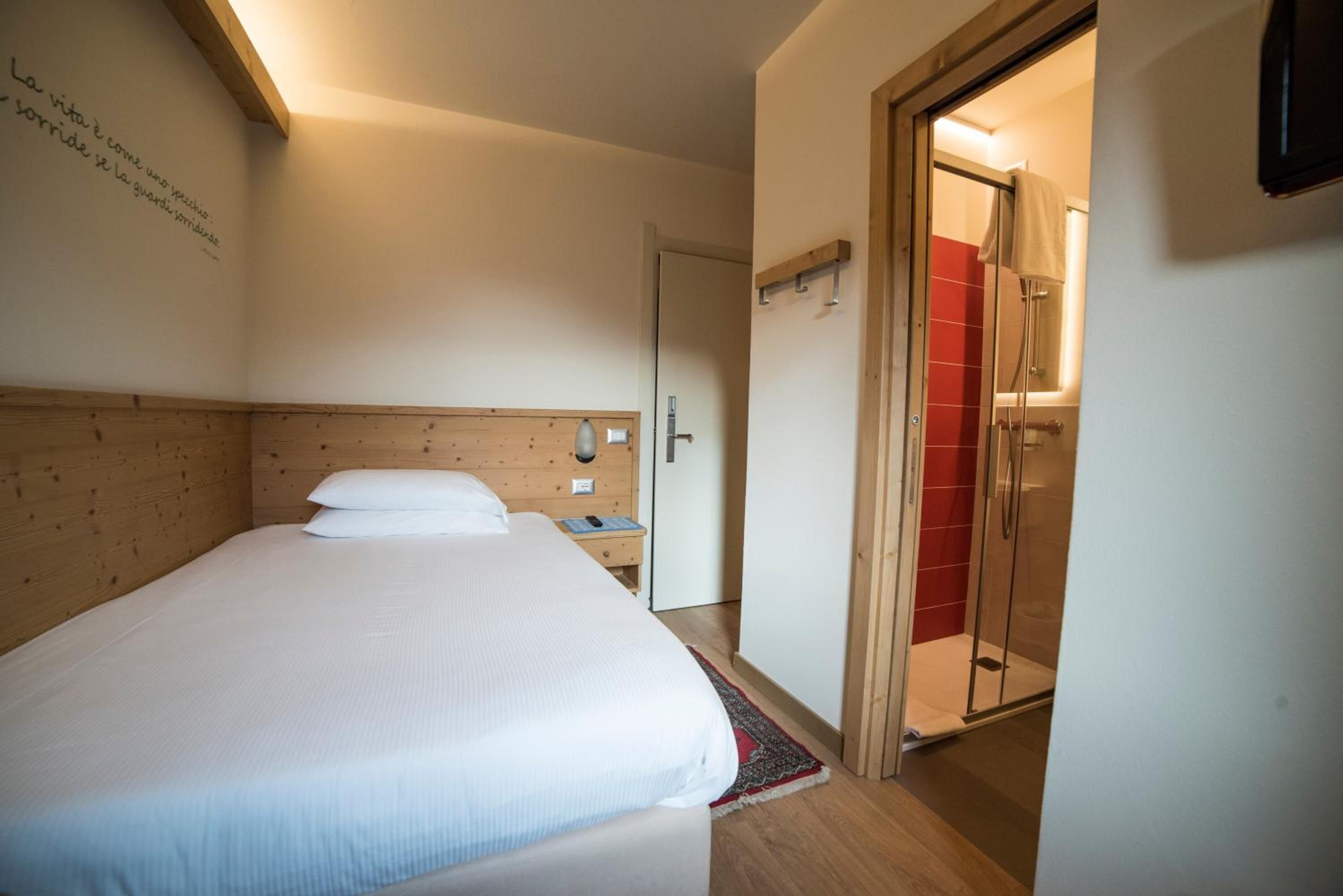 Een slaapkamer met een houten hoofdbord, een bed met wit beddengoed, een nachtkastje en een klein nachttafeltje. Er is een deur naar een badkamer met een douche en een rode betegelde wand.