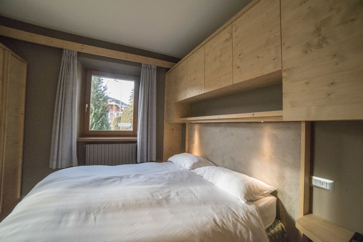 Un dormitorio con cama, cabecera de madera, ventana con cortinas y vista a un edificio.