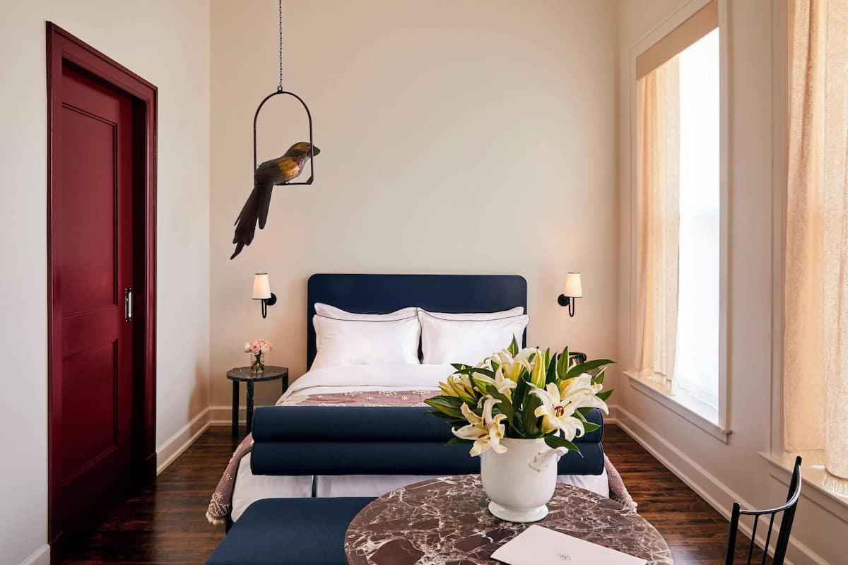 A bedroom with a dark blue bed, white pillows, a small bedside table with a lamp, a vase of flowers, and a hanging bird decoration.