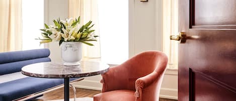 A room with a blue sofa, a round table with a vase of flowers, a pink armchair, and a wooden floor.