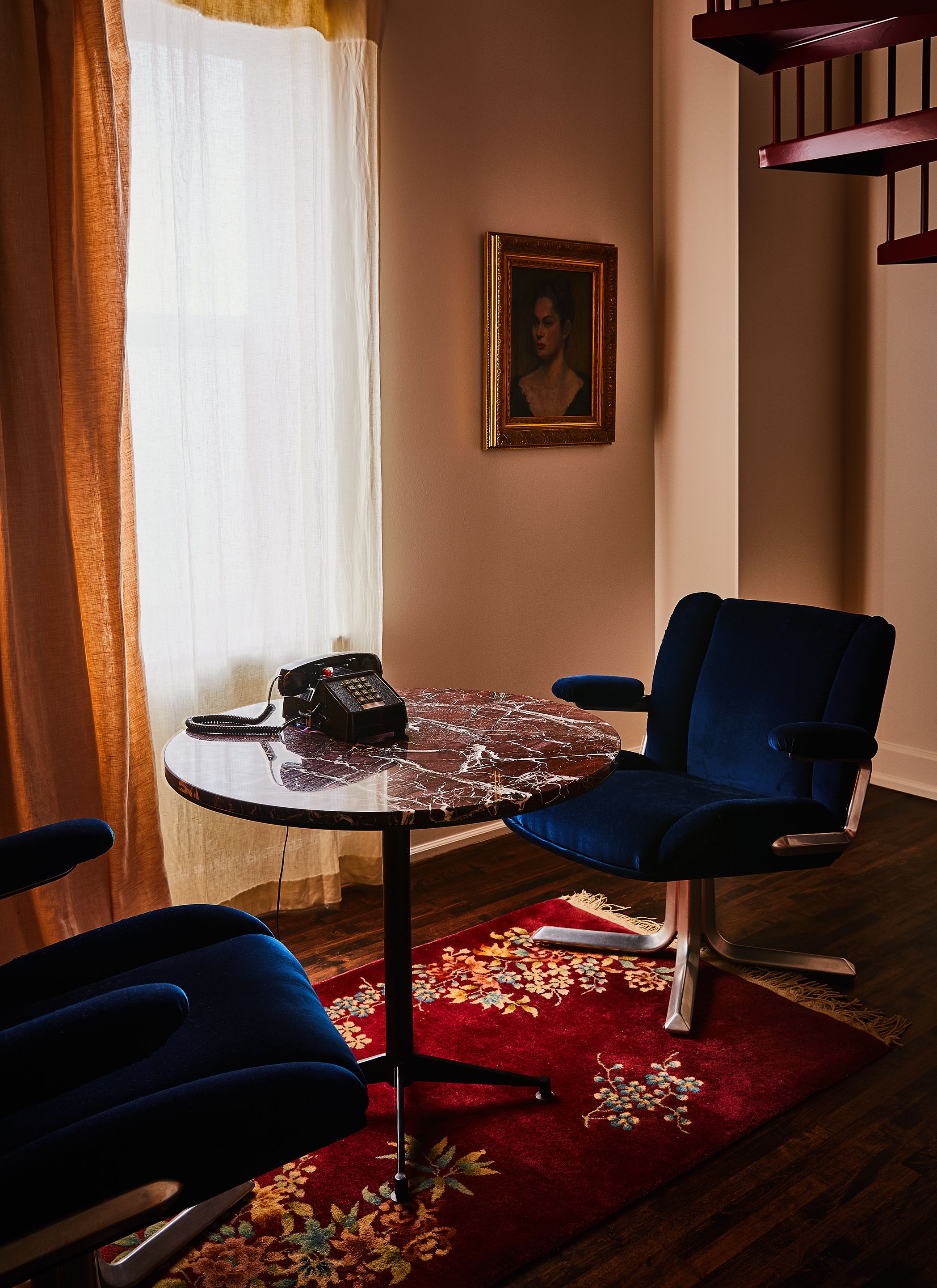 Una habitación con una mesa redonda, dos sillas azules, un tapete floral rojo, un teléfono y un cuadro en la pared.
