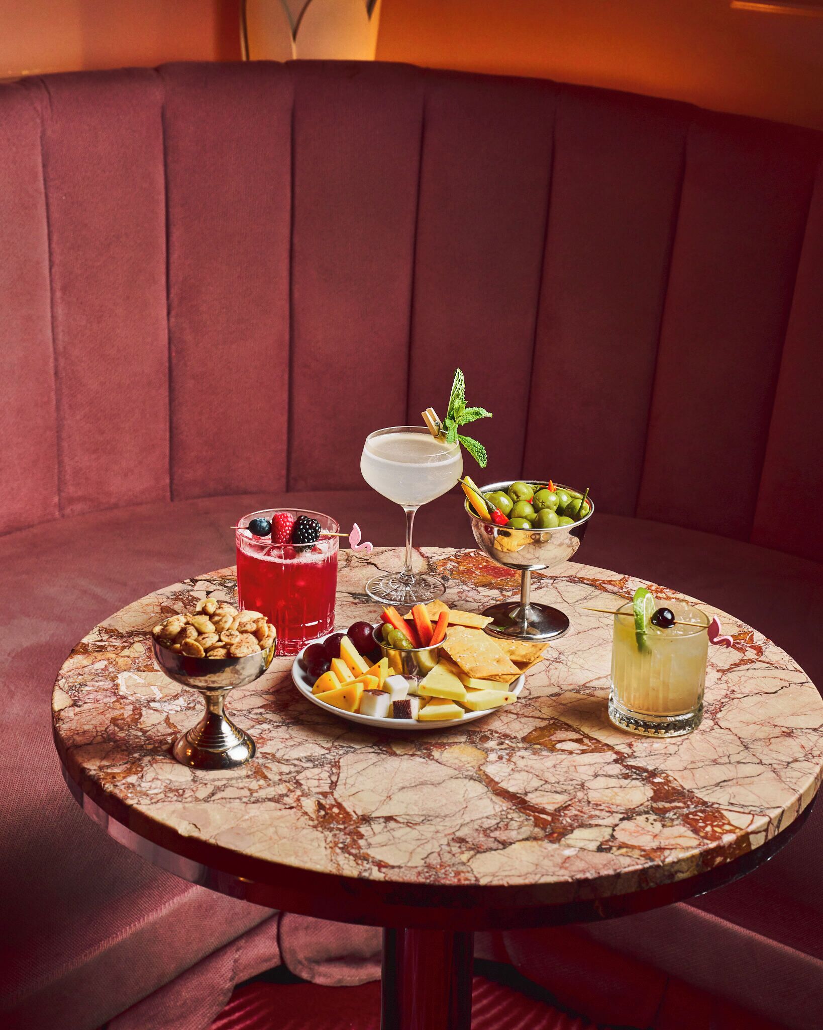 A round marble table with a variety of food and drinks, set on a red upholstered bench.