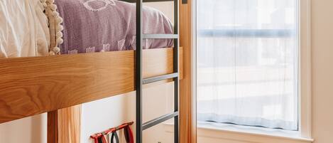 A bunk bed with a desk and chair, a window with curtains, and a small table with a lamp and a cup.