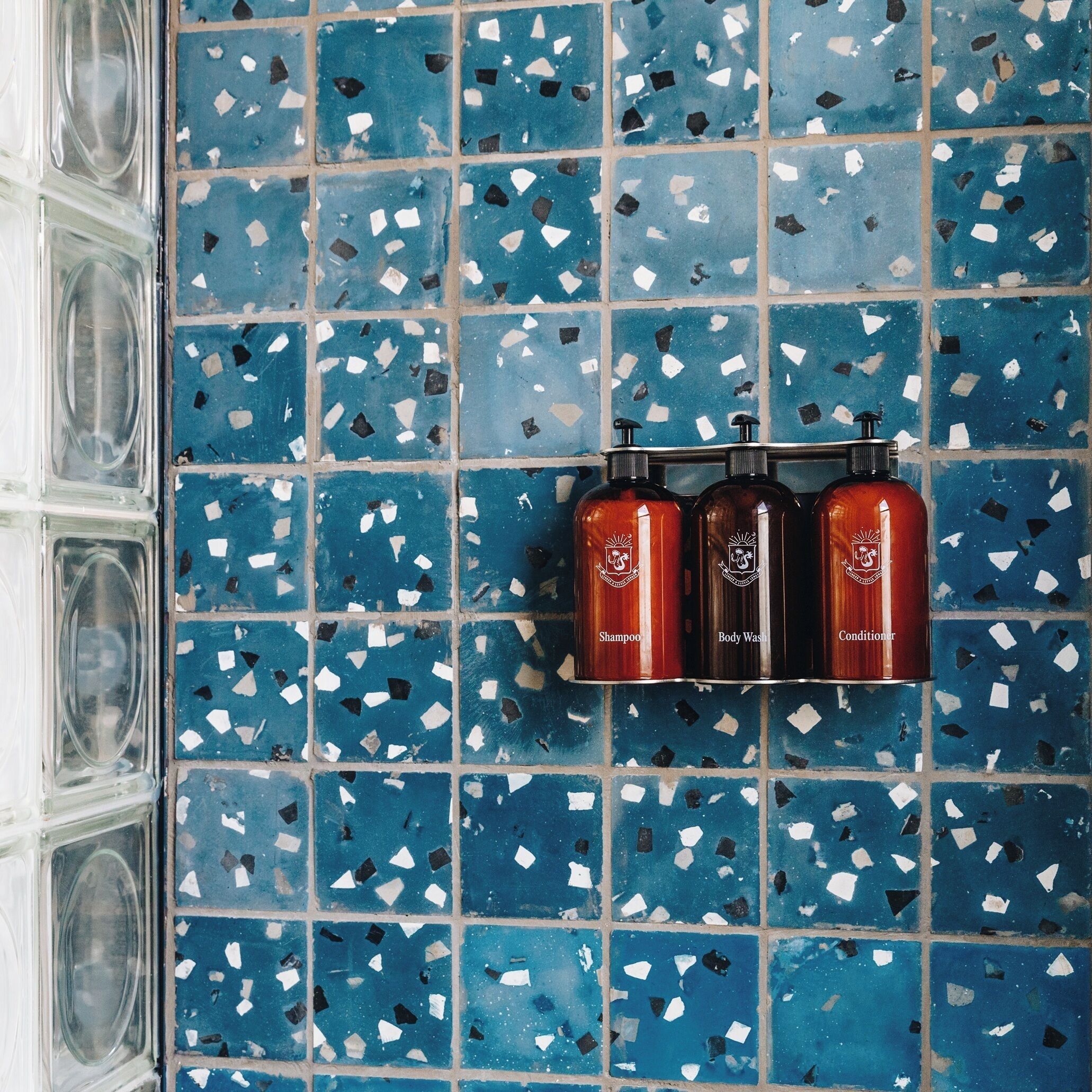 A bathroom with blue mosaic tiles and three bottles of shampoo, body wash, and conditioner.