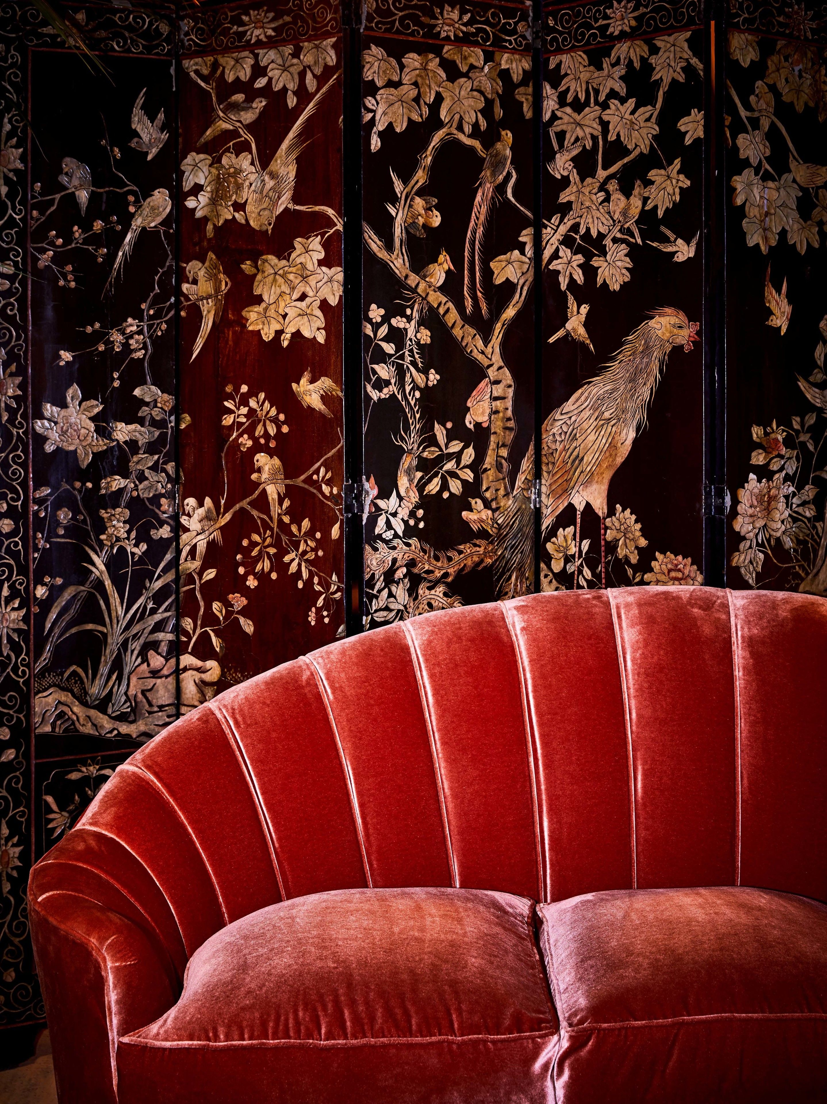 Ein rotes Samtsofa vor einer dekorativen Wandverkleidung mit aufwendigen Vogel- und Blumendarstellungen.