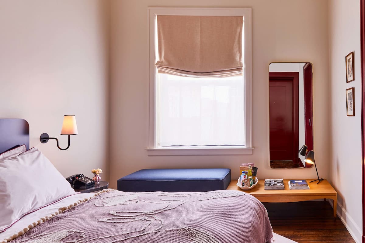 A bedroom with a bed, bedside lamp, window with curtains, bedside table, mirror, and a small bench.