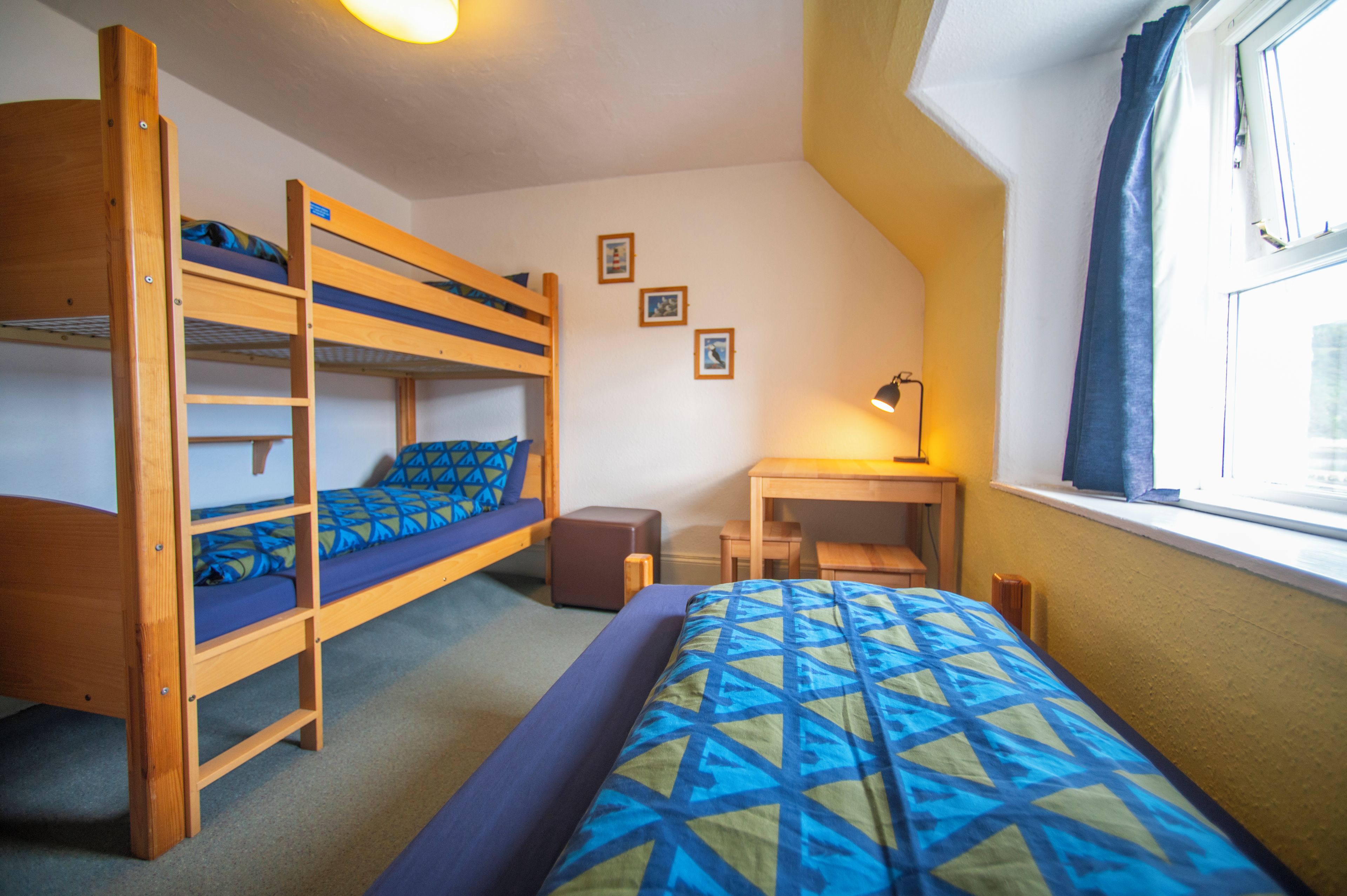 A dormitory room with bunk beds, a desk, a chair, and a window with curtains.