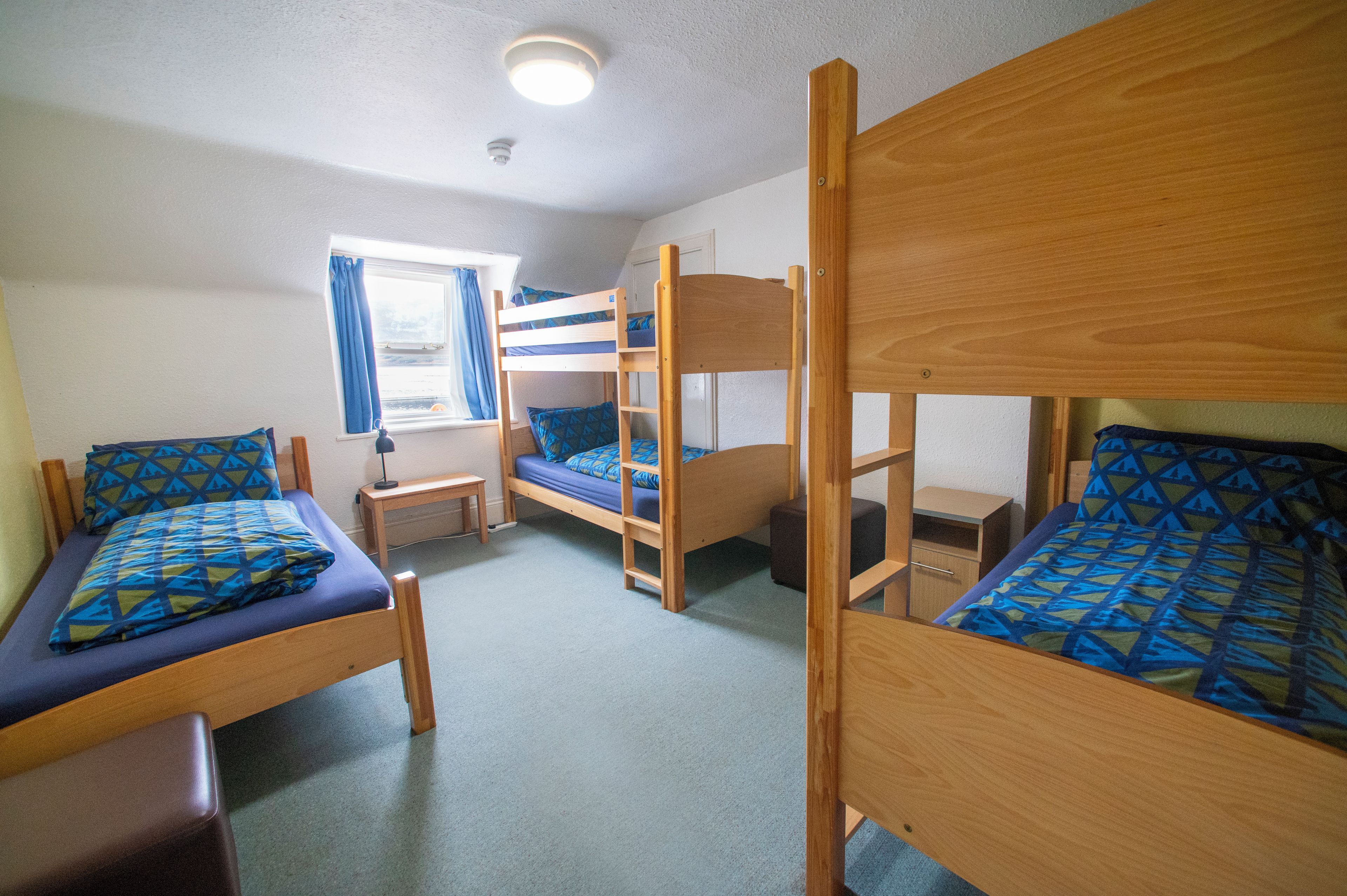 A dormitory room with bunk beds, a small desk, a chair, and a window with curtains.