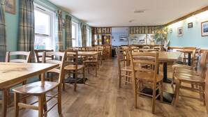 A dining area with wooden tables and chairs, a window with curtains, and a wall with framed pictures. - Ullapool Youth Hostel (Ullapool)