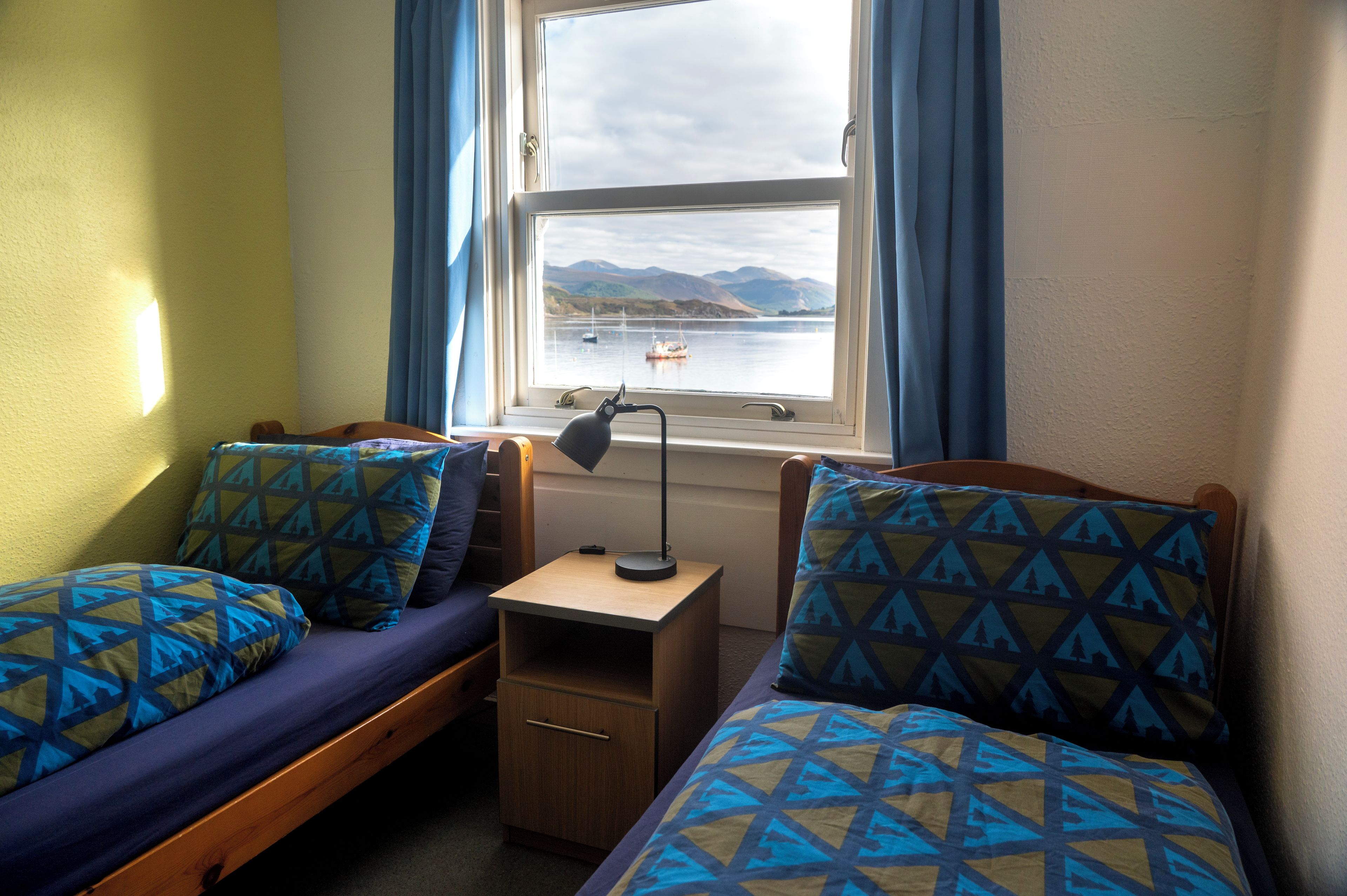Une chambre avec deux lits simples, une petite table de chevet en bois, un bureau avec une lampe et une fenêtre donnant sur un paysage de montagnes et d’eau.