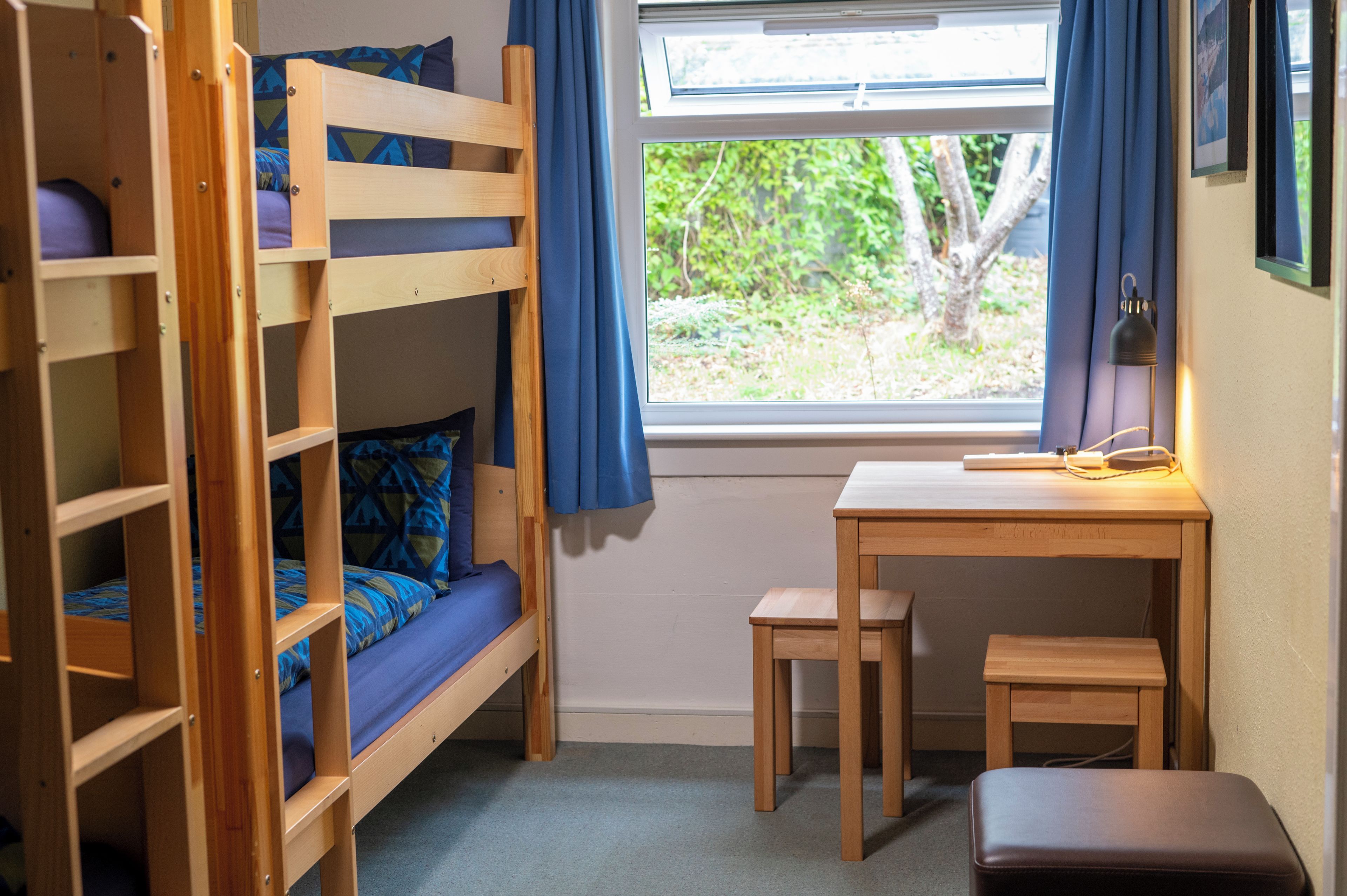 A bunk bed room with a desk and chair, a window with curtains, and a view of greenery outside.