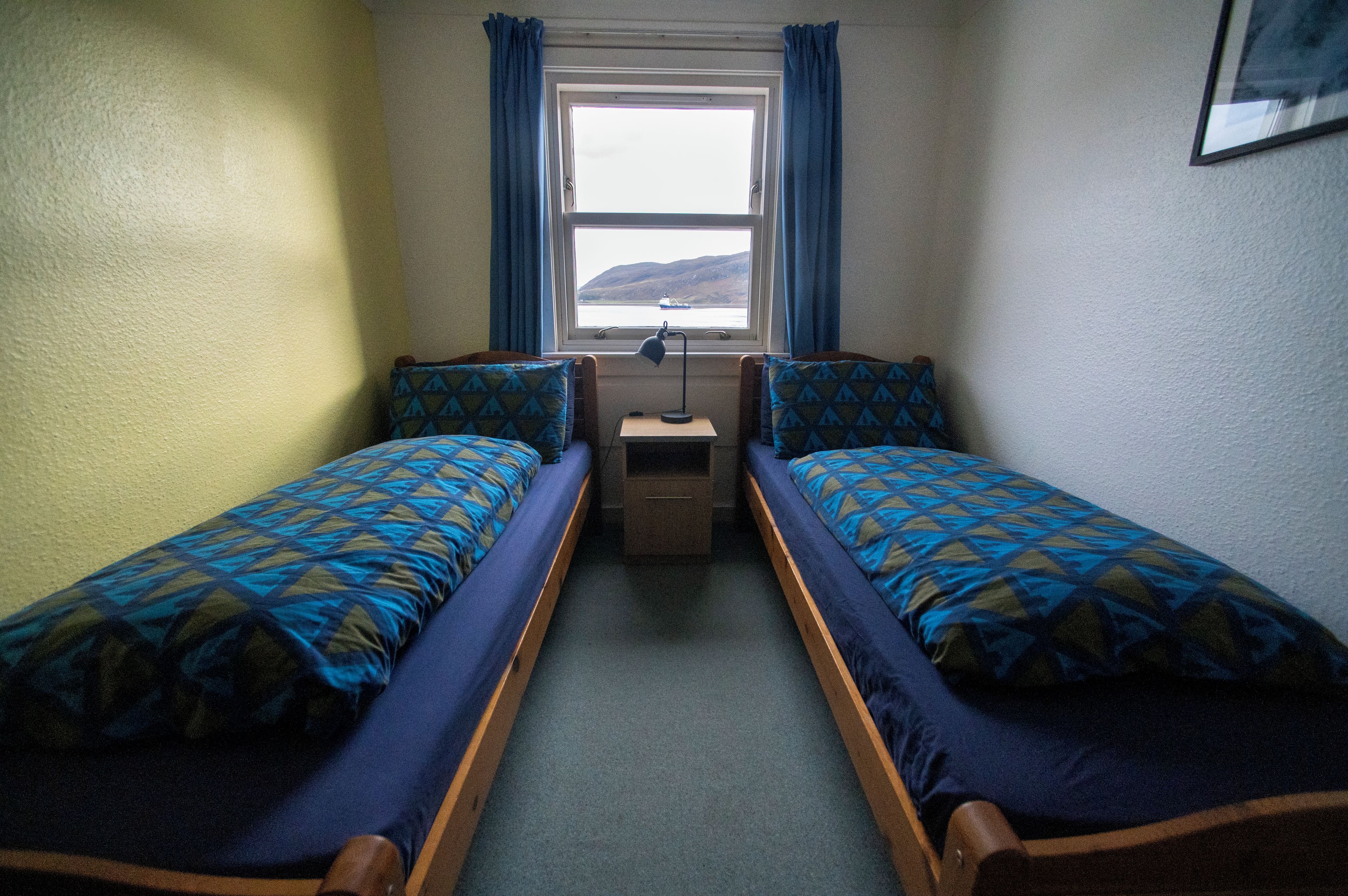 Une chambre avec deux lits simples, une petite table, une lampe et une fenêtre donnant sur l’extérieur.