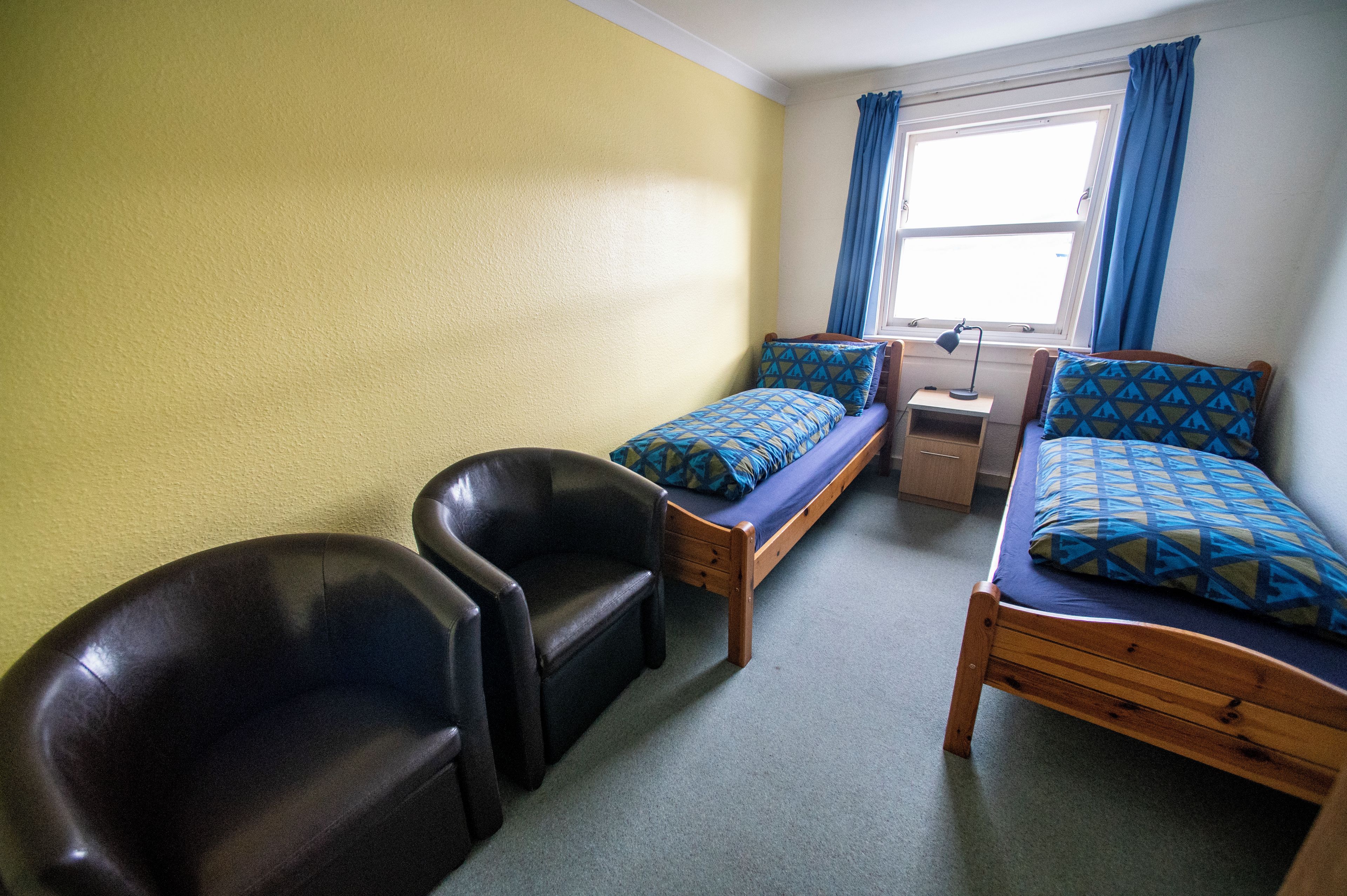 A dormitory room with two single beds, a desk, a chair, and a window with curtains.