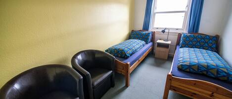 A dormitory room with two single beds, a desk, a chair, and a window with curtains.