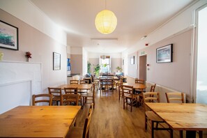A spacious dining area with multiple wooden tables and chairs, a central hanging light fixture, and framed artwork on the walls. - Gairloch Sands Youth Hostel (Gairloch)