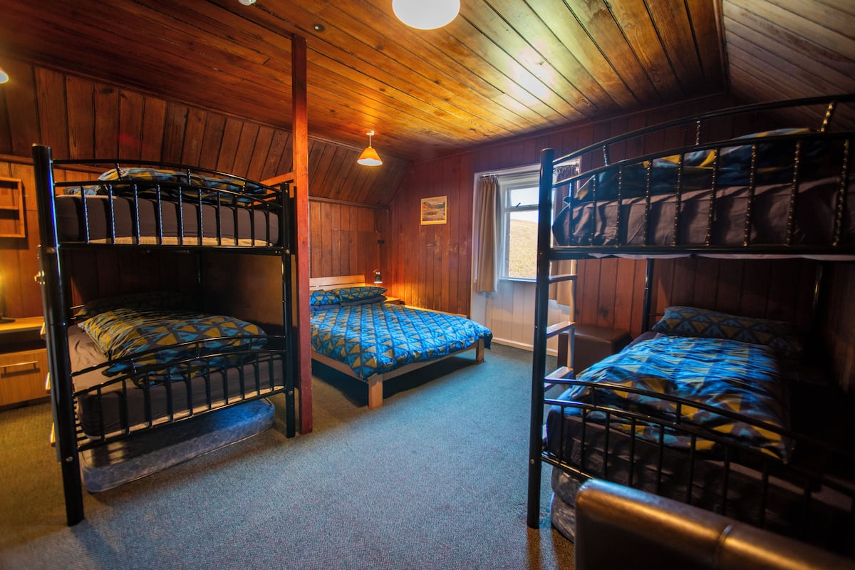 A room with three bunk beds, wooden walls, a window, and a single hanging light fixture.